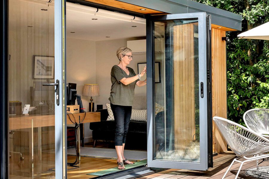 A woman is opening a sliding glass door of a bespoke garden room with a desk, chair, and couch inside. The office is next to a deck with outdoor furniture and plants in the background.