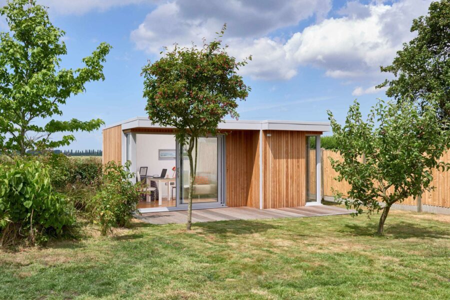 A modern, small wooden house with large glass doors is surrounded by greenery, including trees and a lush lawn, under a partly cloudy sky. This charming setting includes a serene garden room from eDEN Garden Rooms, offering an idyllic retreat.