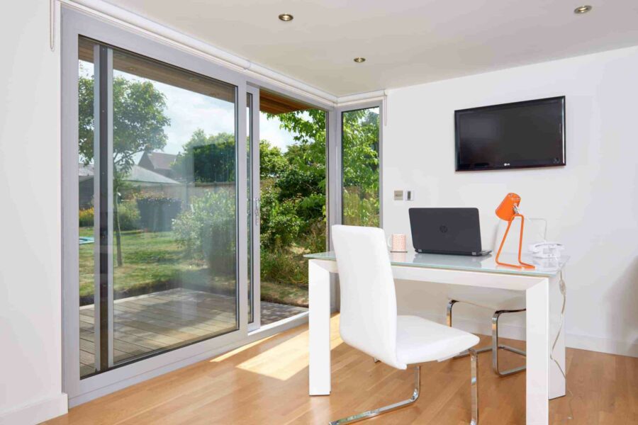 Bright home office with a glass door leading to a garden, featuring a white desk with an orange lamp, beige chair, laptop, and mounted flat-screen TV. For all year round usage, consider these garden room tips to ensure your workspace remains cozy and inviting in any season.