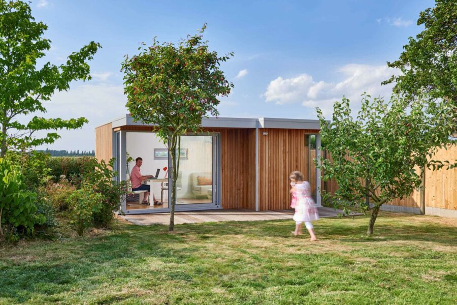 A small modern wooden house with large windows, a person working inside, and a child running on the lawn surrounded by trees and greenery. Designed by eDEN Garden Rooms, this home embodies the innovation that earned it the 2020 Design Award.