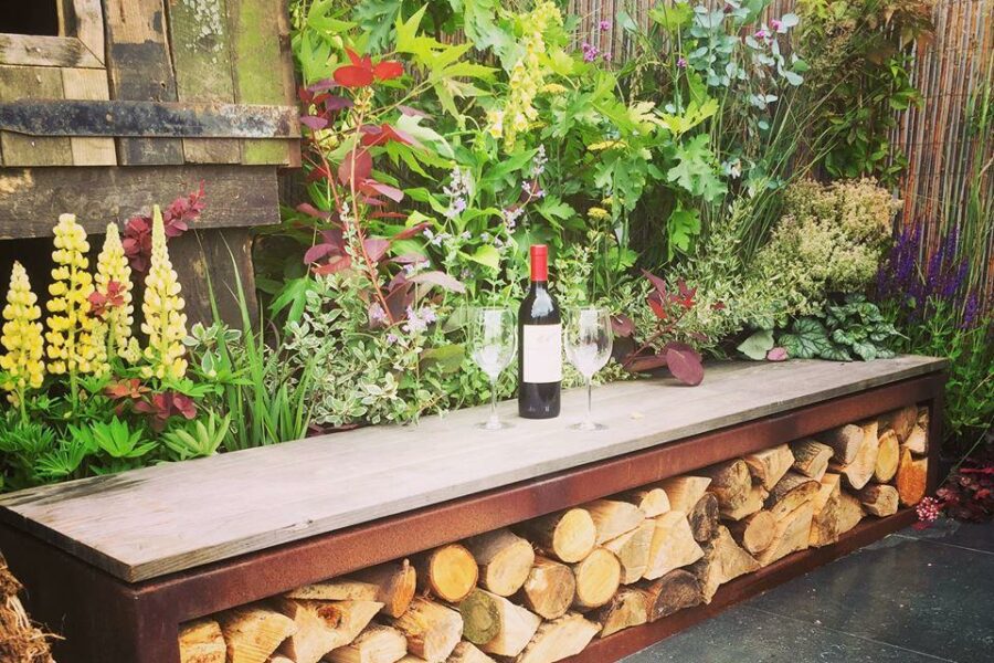 A bench with a compartment filled with firewood is placed in a garden with flowers and plants, perfect for your Outdoor Living space. On the bench are two wine glasses and a bottle of red wine, available from our web shop.