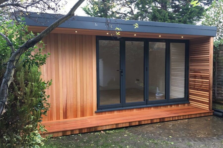 A modern wooden shed with large glass doors, situated in a garden with surrounding trees and shrubs, serves as a multi-functional studio for various creative pursuits.