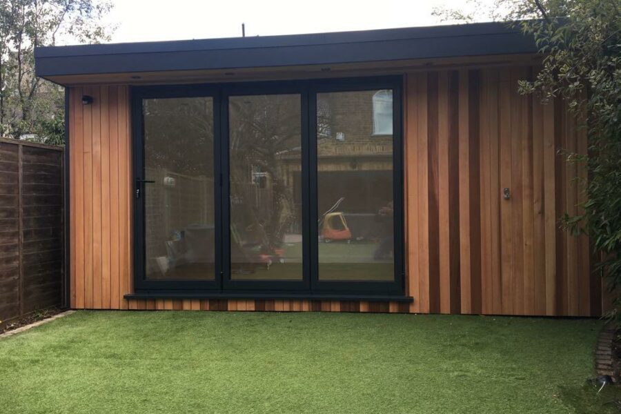 A small, modern shed with large glass doors and wooden paneling, the eDEN is situated in a Richmond garden with artificial grass and surrounded by a wooden fence. This resourceful room offers both style and functionality.