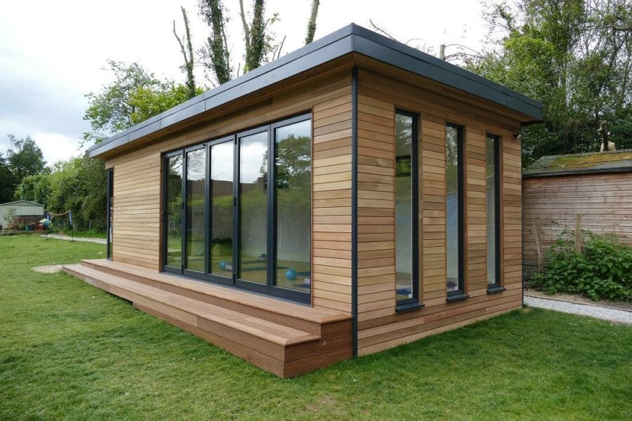 A modern, rectangular one-story wooden building with large glass windows, surrounded by a grassy area and trees, epitomizing the high-quality design of eDEN insulated garden rooms.