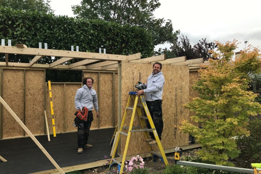 Two men working on constructing a wooden structure; one man stands on a ladder using a drill, while the other holds a chainsaw. The area is surrounded by greenery, reflecting the collaborative spirit of the growing eDEN team.