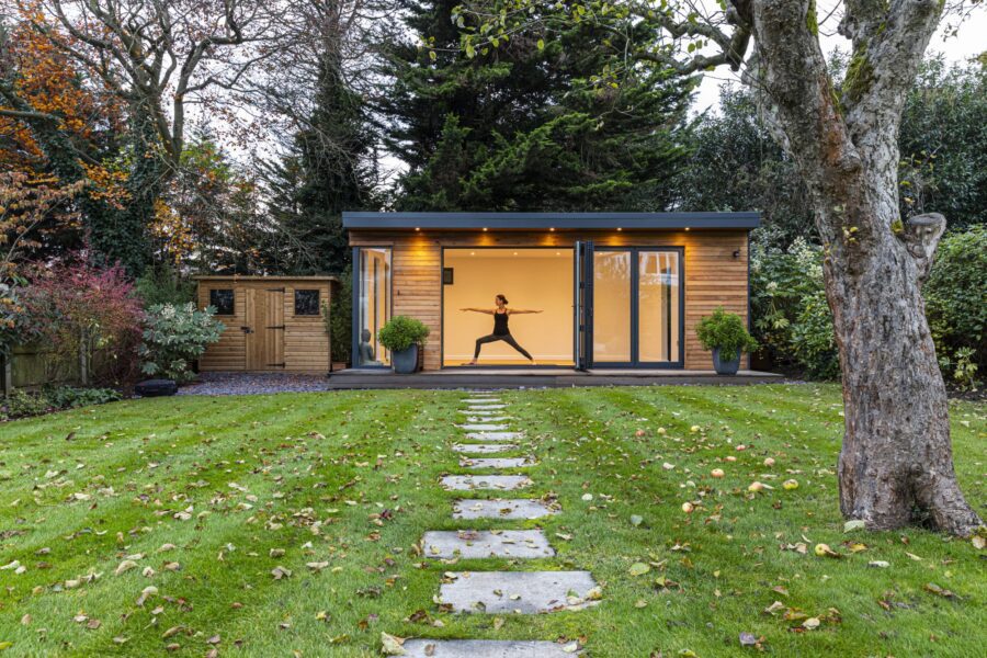 A person performs a yoga pose inside a bespoke yoga studio in Edgware, surrounded by a well-maintained lawn with stepping stones, trees, and a wooden shed in the background.