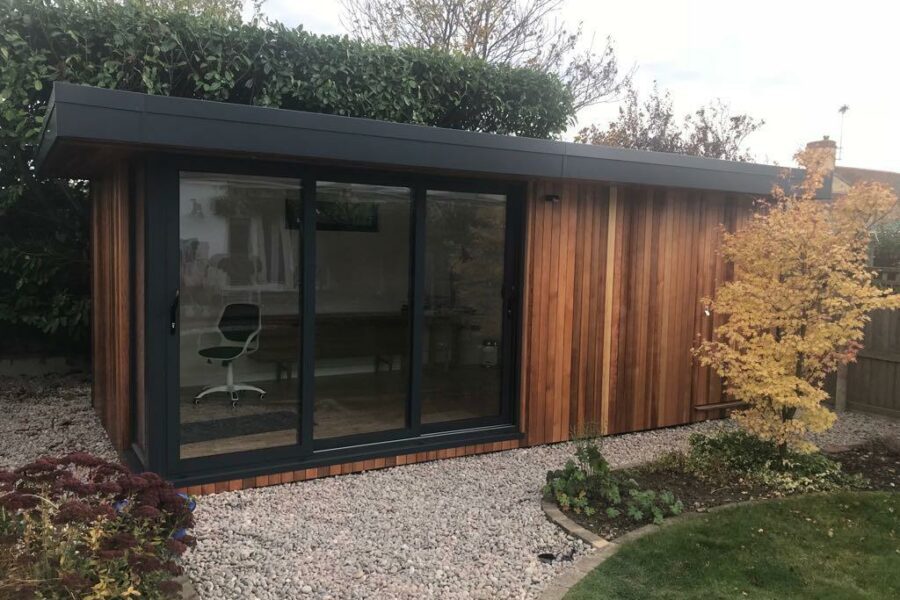 A modern, multi-functional garden office with large glass sliding doors and a wood-paneled exterior is surrounded by gravel and landscaped garden features, including a small tree on the right. Perfect for creating your own Whitstable-inspired retreat.