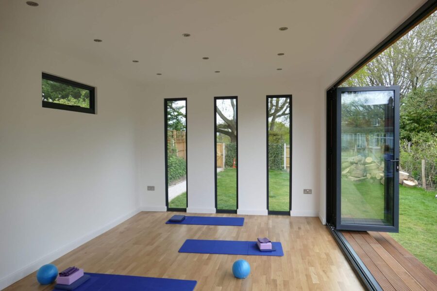 A bright bespoke garden room with wooden floors features yoga mats, blocks, and exercise balls. Large vertical windows and an open door reveal the serene garden outside.