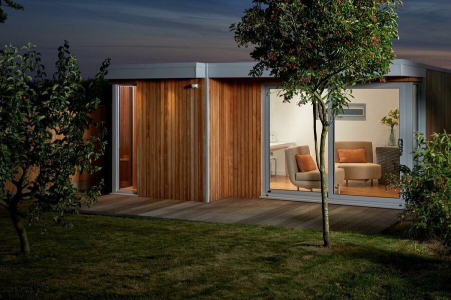 A modern wooden garden room with large glass doors, illuminated at dusk, invites you to pause. The interior shows a cozy seating area with two armchairs and a small table. Trees surround the structure, making it an ideal retreat for garden room projects or relaxation.