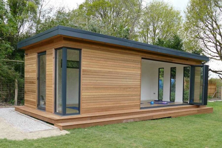 A modern, rectangular wooden cabin with large glass windows and a flat roof, situated on a grassy area and surrounded by trees, serves as a versatile room perfect for any home.