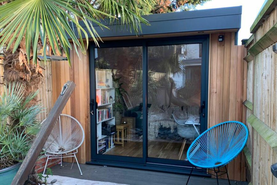 A small wooden garden room in a compact space with large glass sliding doors, surrounded by greenery. Inside, it is equipped with a desk, bookshelf, and chair. Blue and black chairs are positioned on the outside deck.