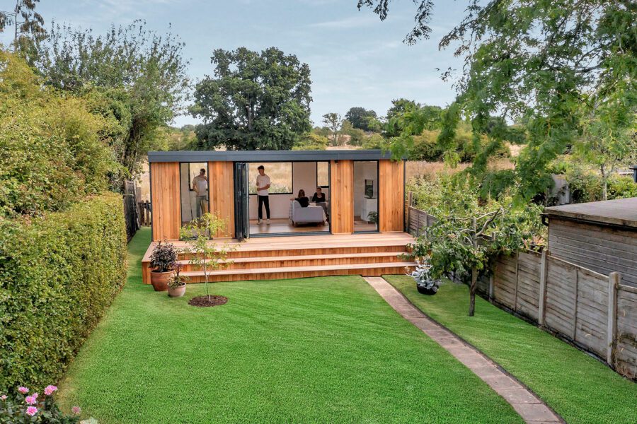 A modern wooden backyard house with large windows, a patio, and a well-maintained lawn, surrounded by greenery and adjacent to a wooden fence and small shed. These insulated garden rooms offer comfort year-round.