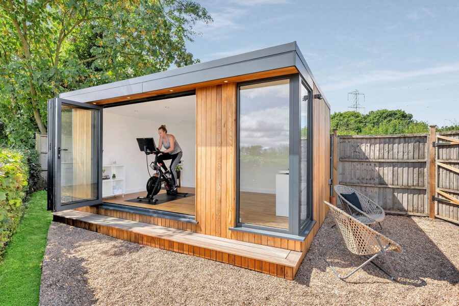 A person exercises on an indoor stationary bike in a modern insulated garden room with large glass doors open to a yard with a hedge, fence, and chair.