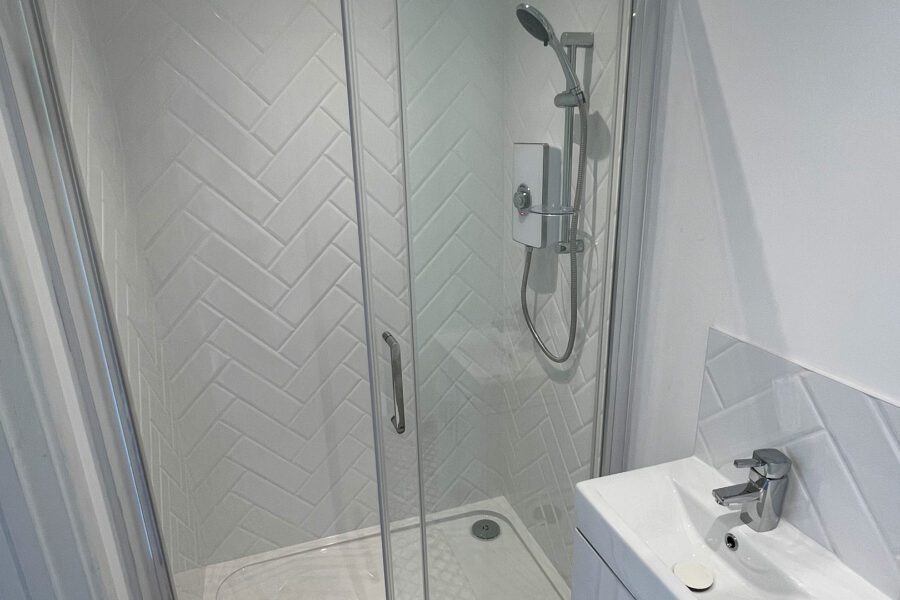 A modern shower cubicle with white herringbone tile walls, a glass door, and a wall-mounted showerhead. The bathroom also features a small white sink with a faucet and a bar of soap to the right, providing both functionality and aesthetic benefits.