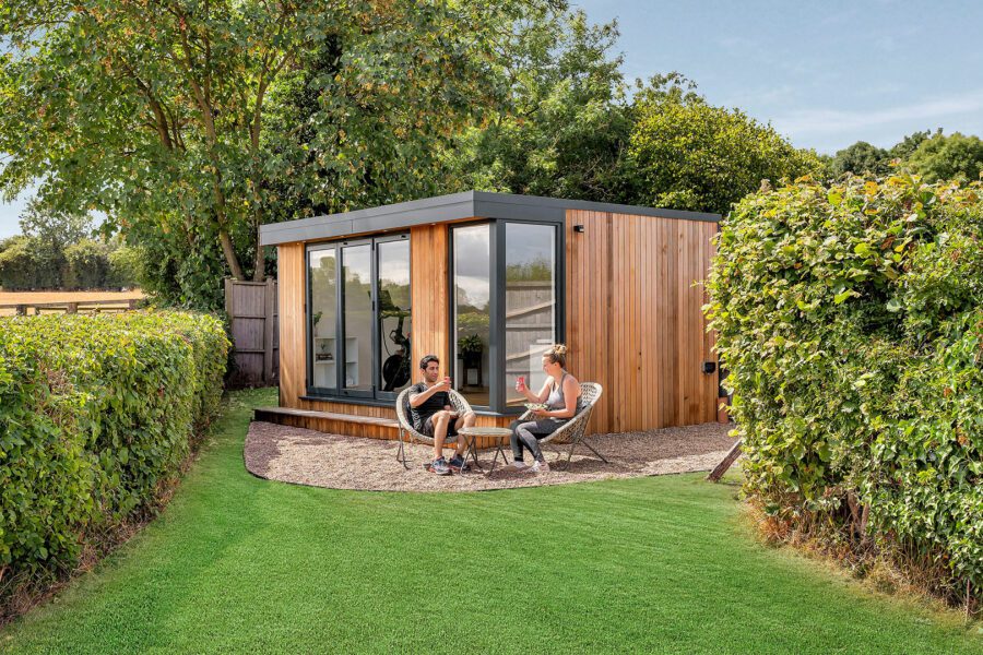 Two people sit and converse on a small patio in front of a modern wooden cabin, surrounded by green hedges and trees. This serene setting perfectly showcases the charm of insulated garden rooms.