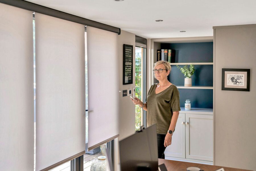 A person adjusts window blinds using a remote control in a modern, well-lit insulated garden room with bookshelves and minimalist decor.