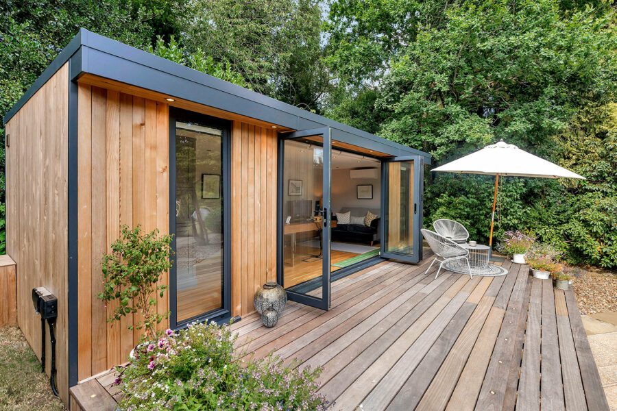 A modern tiny house with a wooden exterior, large glass doors, and a patio featuring outdoor furniture and an umbrella, surrounded by lush greenery. This versatile space can double as one of those ideal garden offices for remote work amidst nature.