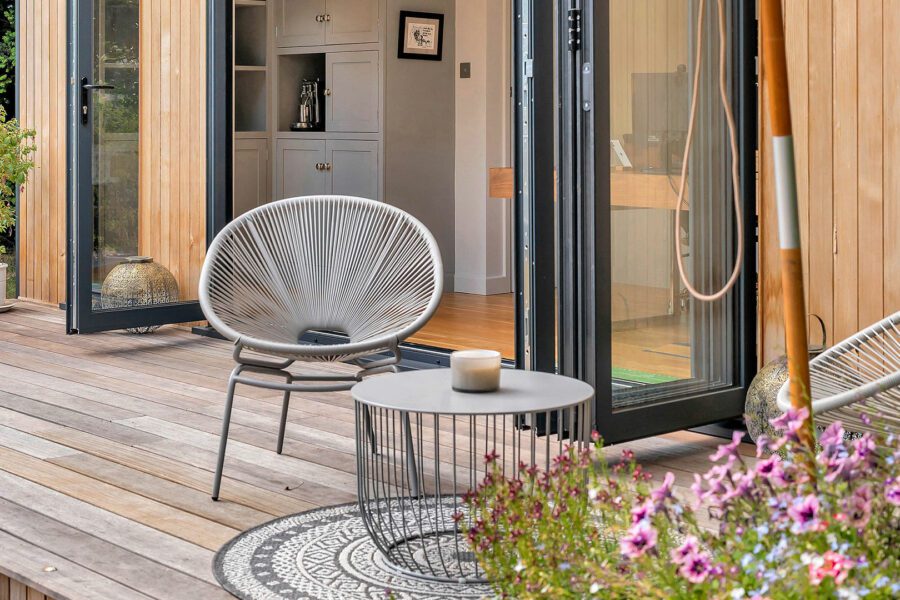 A patio with a circular table, candle, and two modern chairs. The space features wooden flooring, a potted plant, and an open door leading to an interior room with cabinets and decor—perfect for incorporating garden room ideas.