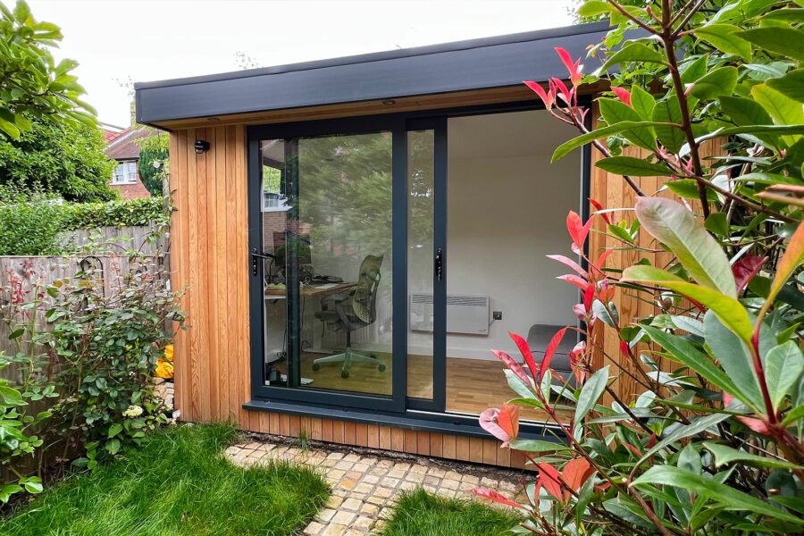 Wooden garden studio office with large glass doors surrounded by greenery and plants. Inside, a desk and office chair are visible through the glass.