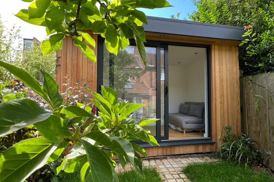 A small wooden garden office with a glass sliding door, partially surrounded by green plants and trees. Inside, there is a gray sofa. A brick path leads to the entrance—one of those charming small garden rooms that blend seamlessly with nature.