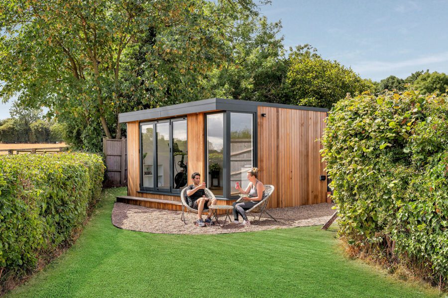 Two people sit at a small table in front of a modern, wood-paneled tiny house, surrounded by green hedges and a manicured lawn, enjoying their sunny garden room retreat.