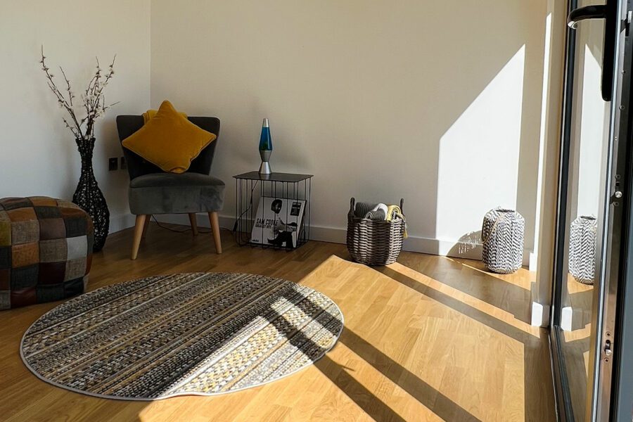 A sunlit room with a wooden floor features a small round rug, a gray chair with a yellow cushion, a colorful pouf, and baskets along the wall—perfect for designing the interior of your garden office. A small table with a lamp and magazines completes this cozy setup.