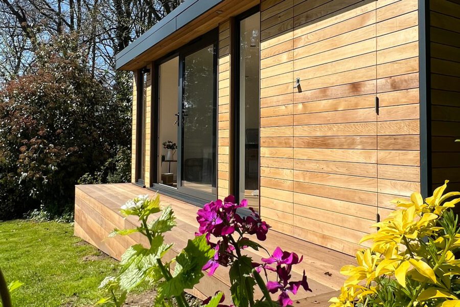 A small wooden house with large sliding glass doors and a wooden deck, surrounded by colorful flowers and lush greenery, serves as the perfect garden studio.