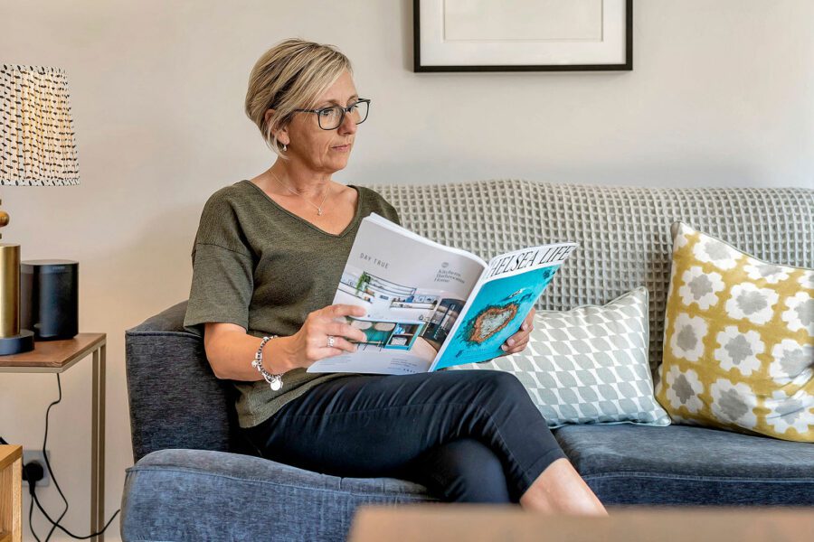 A woman with short hair and glasses sits on a couch, reading a magazine about the benefits of an insulated garden office. She is wearing a green top and black pants.