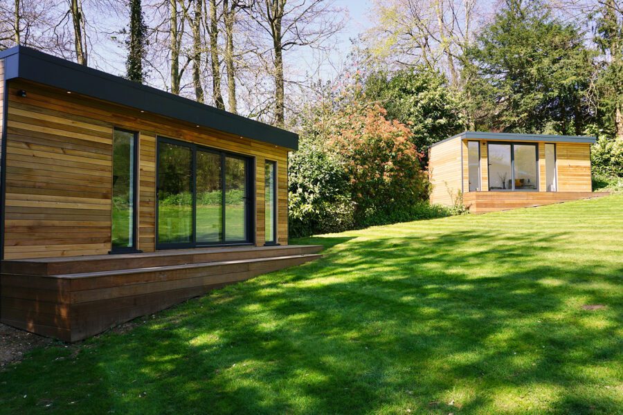 Two modern, small wooden garden studios with large glass doors sit on a well-maintained grassy lawn surrounded by trees and shrubs.