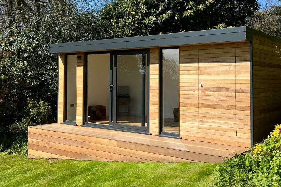 A contemporary wooden garden shed with large glass door and windows, integrating a small garden room, situated on a well-maintained lawn surrounded by greenery.