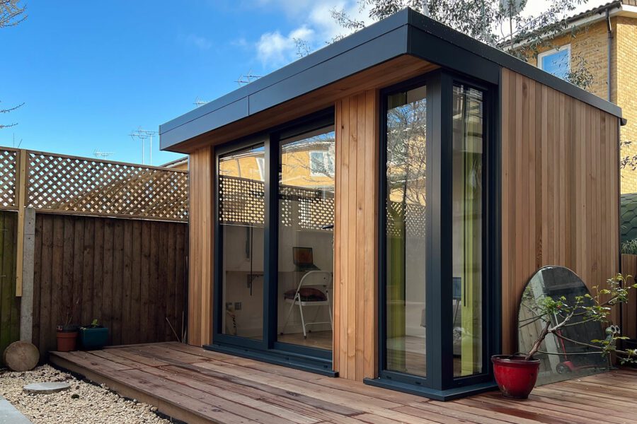 A modern wooden garden studio with large glass doors and windows, situated on a wooden deck. The surrounding area features a wooden fence and potted plants, with a blue sky and neighboring buildings in the background.