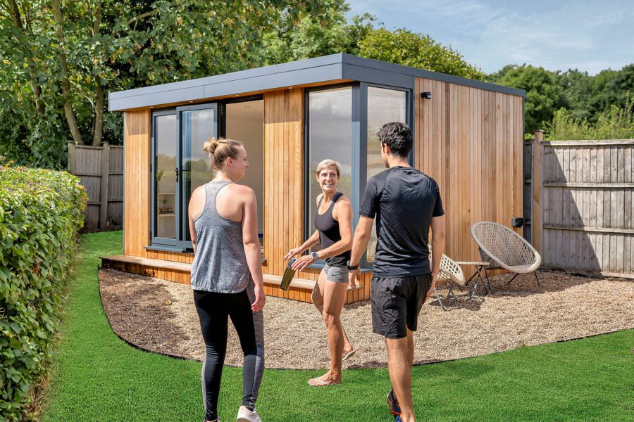 Three people stand and talk near a modern wooden garden room with large glass doors, possibly repurposed as a garden gym. The area features green artificial grass, a wooden fence, and trees in the background.