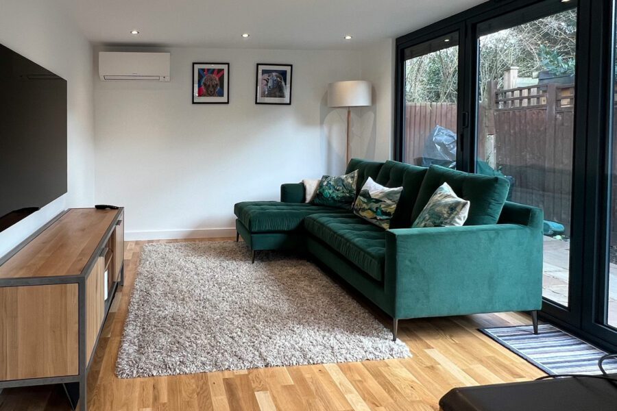 A modern living room features a green sectional sofa, flat-screen TV on a wooden console, a tall lamp, two framed pictures on the wall, and a large sliding glass door opening to an outdoor area with views of an insulated garden office.