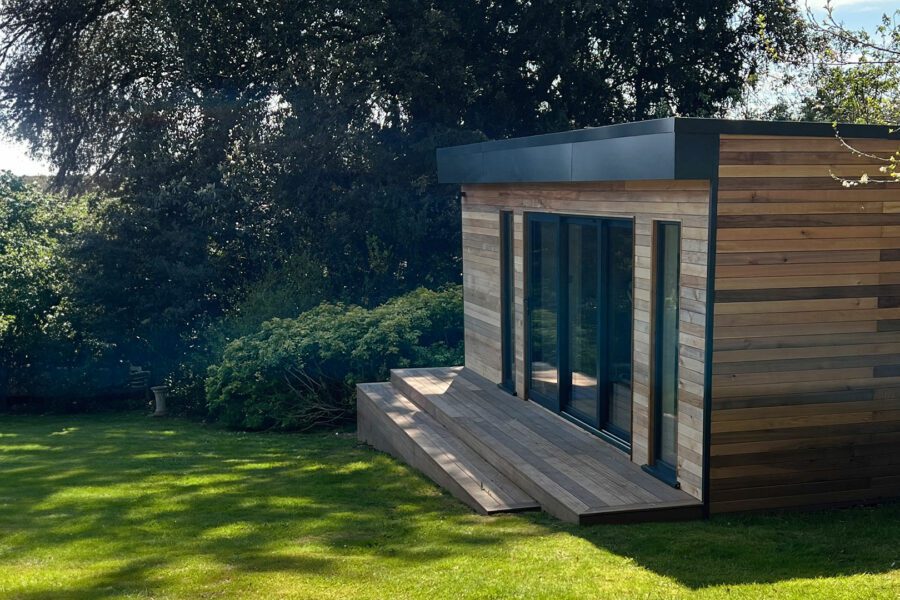 A modern wooden garden shed with large glass doors sits on a well-kept lawn surrounded by lush greenery and trees, offering the perfect setting for stylish garden rooms with bathrooms.