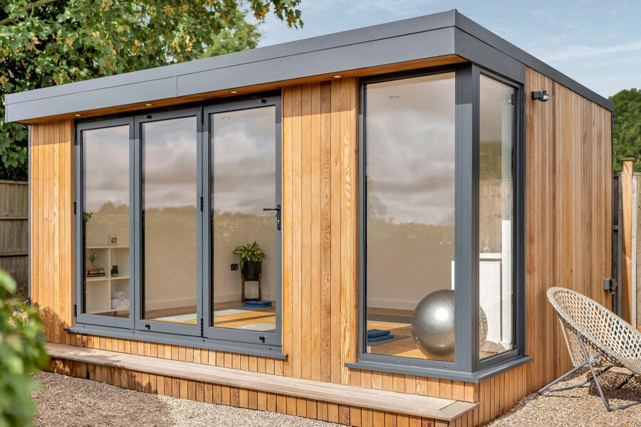 A modern, small wooden house with large glass doors and windows, situated in a backyard with trees, a wooden fence, and adjacent small garden rooms. A wicker chair and exercise equipment are visible inside.