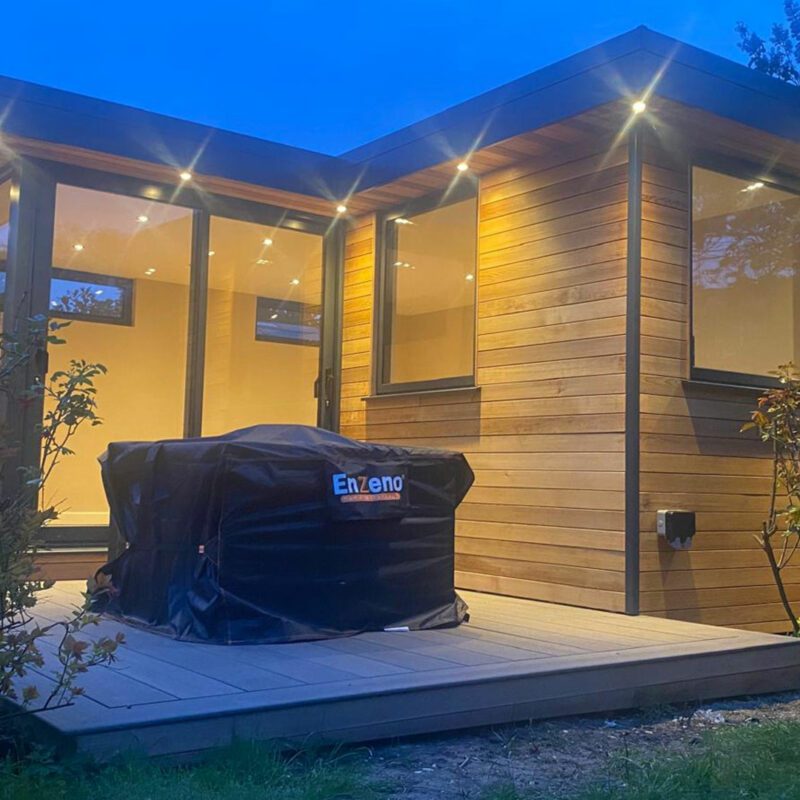 A modern, garden room with large windows and a wooden exterior is beautifully illuminated at dusk. In the foreground, a barbecue grill covered with a black cover sits on the deck, completing the serene outdoor setting.