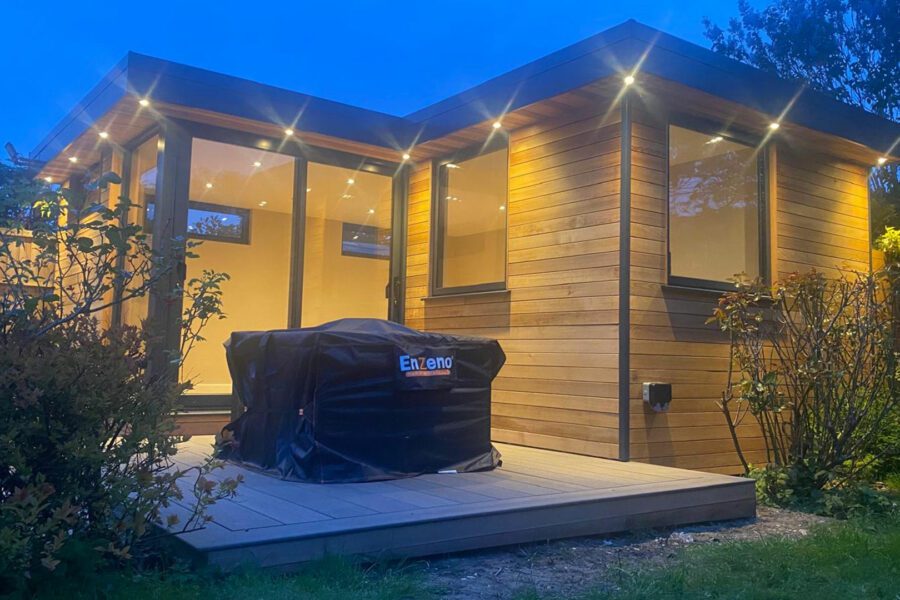 A modern, garden room with large windows and a wooden exterior is beautifully illuminated at dusk. In the foreground, a barbecue grill covered with a black cover sits on the deck, completing the serene outdoor setting.