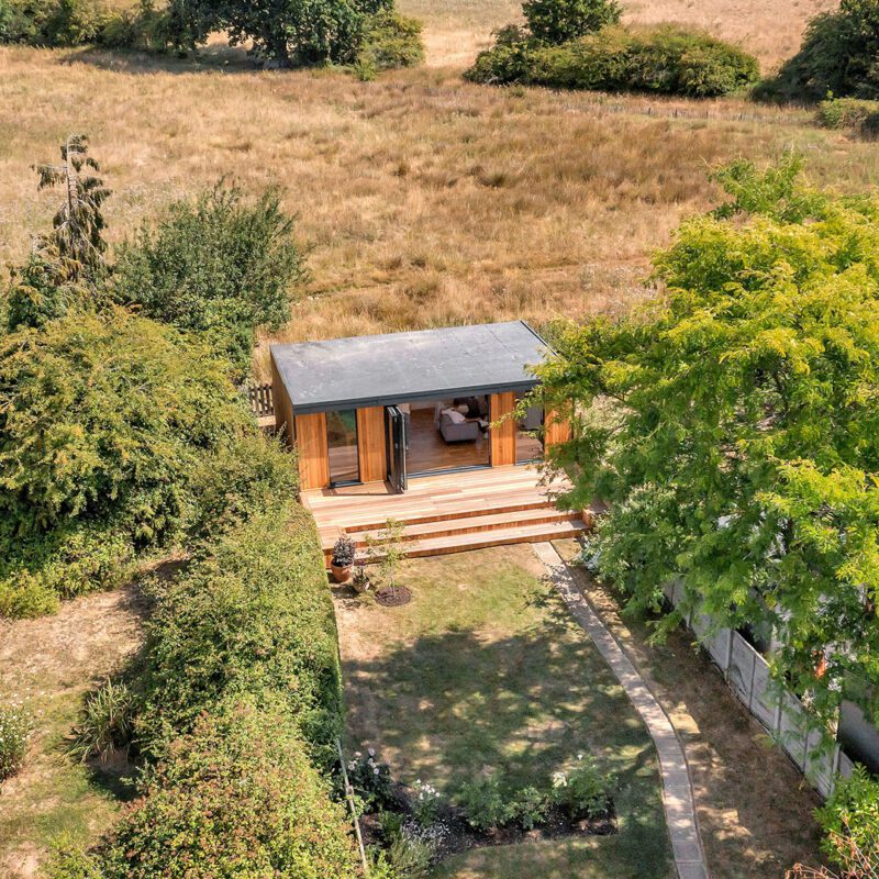 Aerial view of a small, modern cabin with a patio, surrounded by trees and greenery in an open field. A pathway leads from the cabin to the fenced garden studio area.