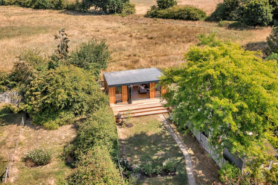 Aerial view of a small, modern cabin with a patio, surrounded by trees and greenery in an open field. A pathway leads from the cabin to the fenced garden studio area.