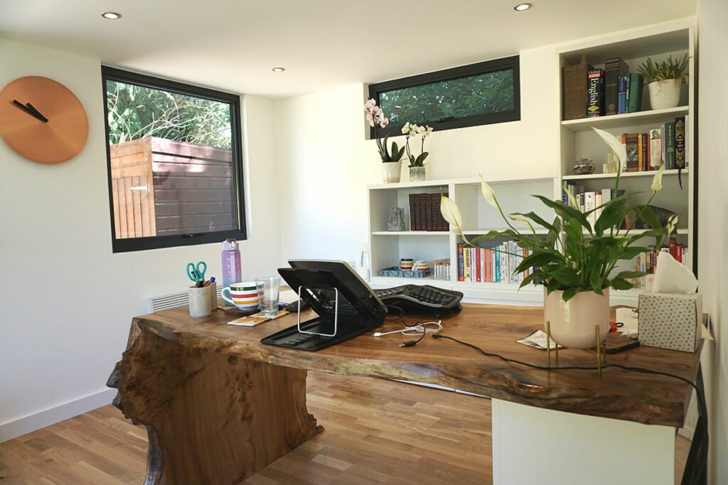 A home office with a wooden desk, computer, plants, and bookshelves. A large window and wall clock are on the left, and shelves with various items are on the right, creating an ambiance similar to an insulated garden office.