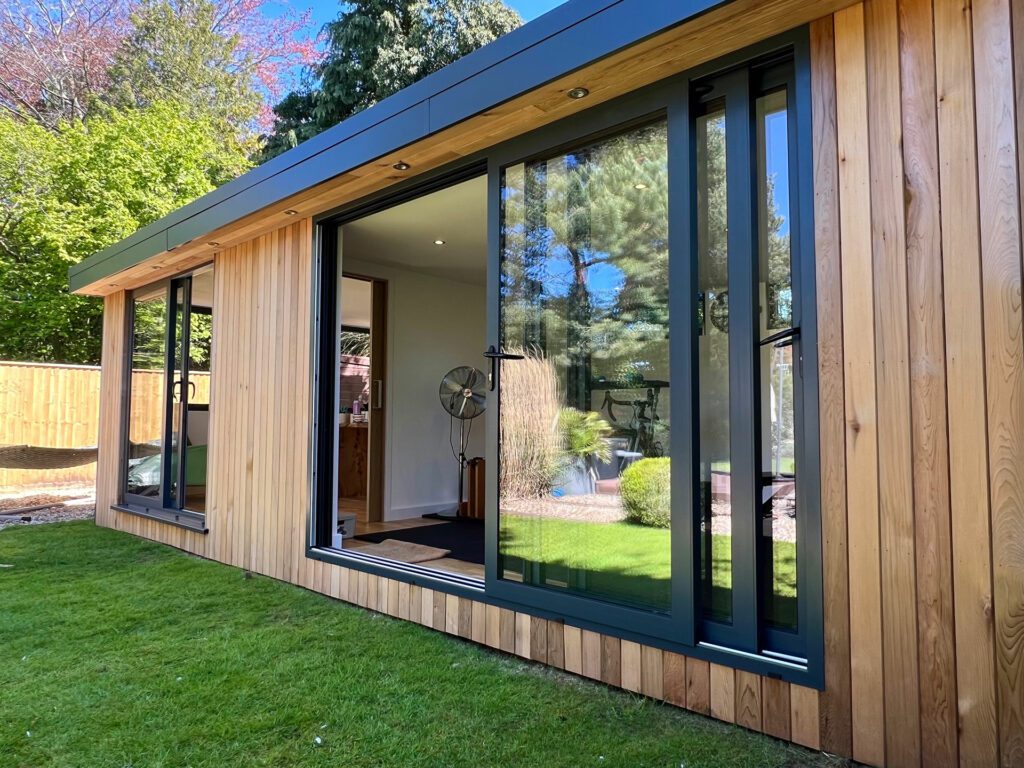 A modern wooden garden room with large sliding glass doors and windows, designed as an insulated garden office, is surrounded by a well-kept lawn and trees in the background.