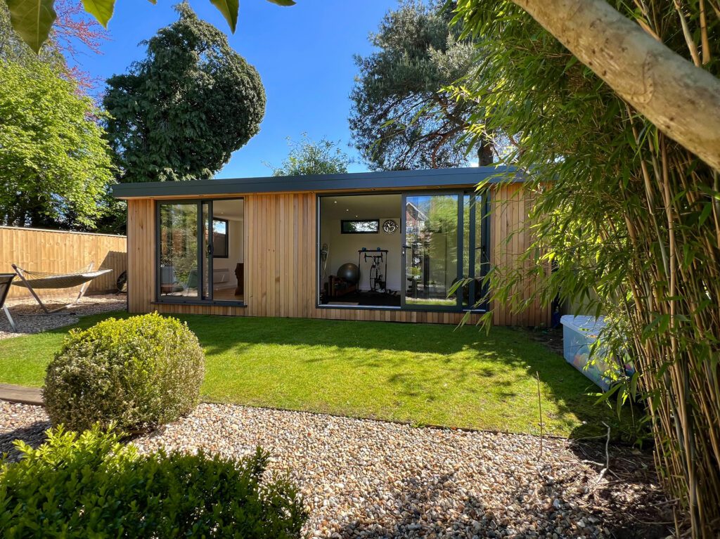 Modern garden office with a wooden exterior, large windows, and a door, set in a landscaped garden with a lawn, gravel paths, trees, and a hammock to the left. Easily convertible into a garden gym for fitness enthusiasts seeking convenience and tranquility.