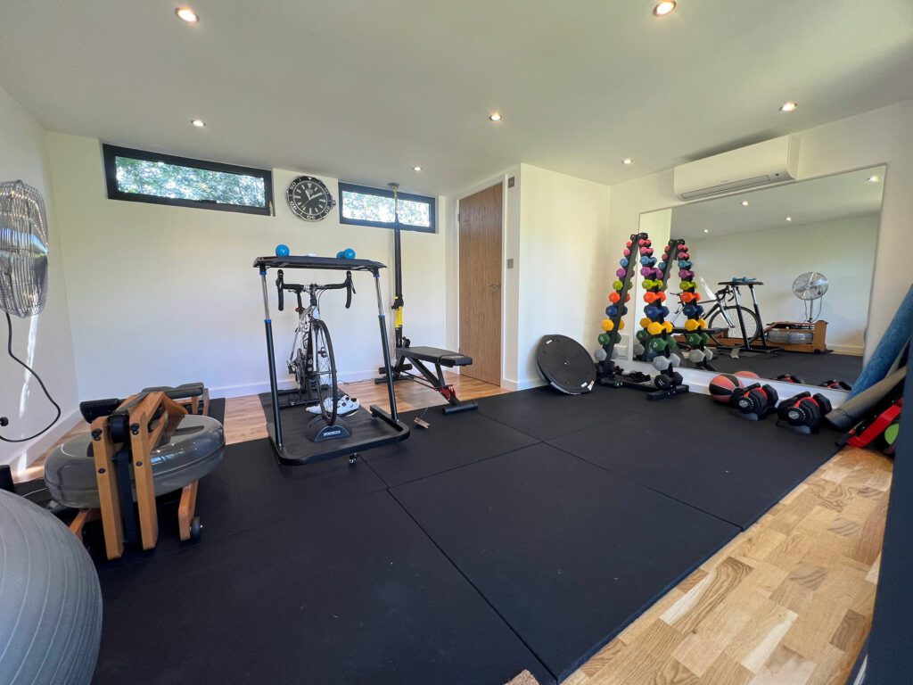 A garden gym featuring various equipment including a stationary bike, free weights, resistance bands, and exercise balls on black mat flooring with mirrors on one wall.