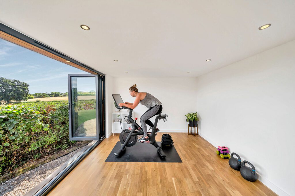 A person exercises on a stationary bike in a bright room with an open glass door revealing a green outdoor landscape, creating the feel of a garden gym. The room has wooden floors, kettlebells, a plant, and small hand weights.