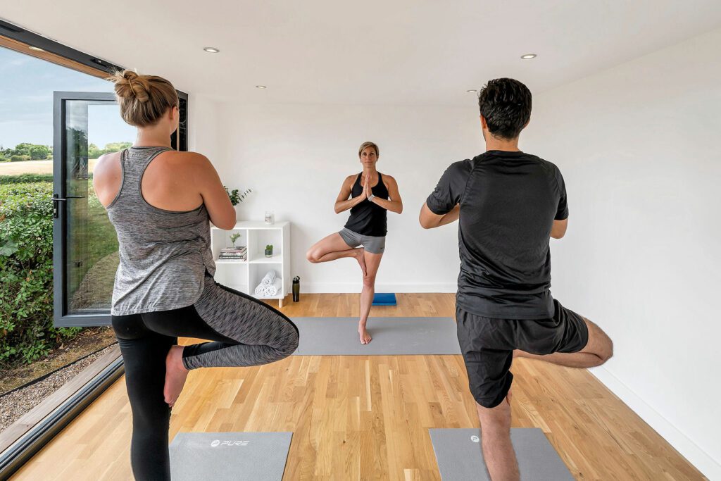 Three people are practicing yoga in a bright garden gym, each balancing on one leg in the tree pose, with an instructor in front guiding them.