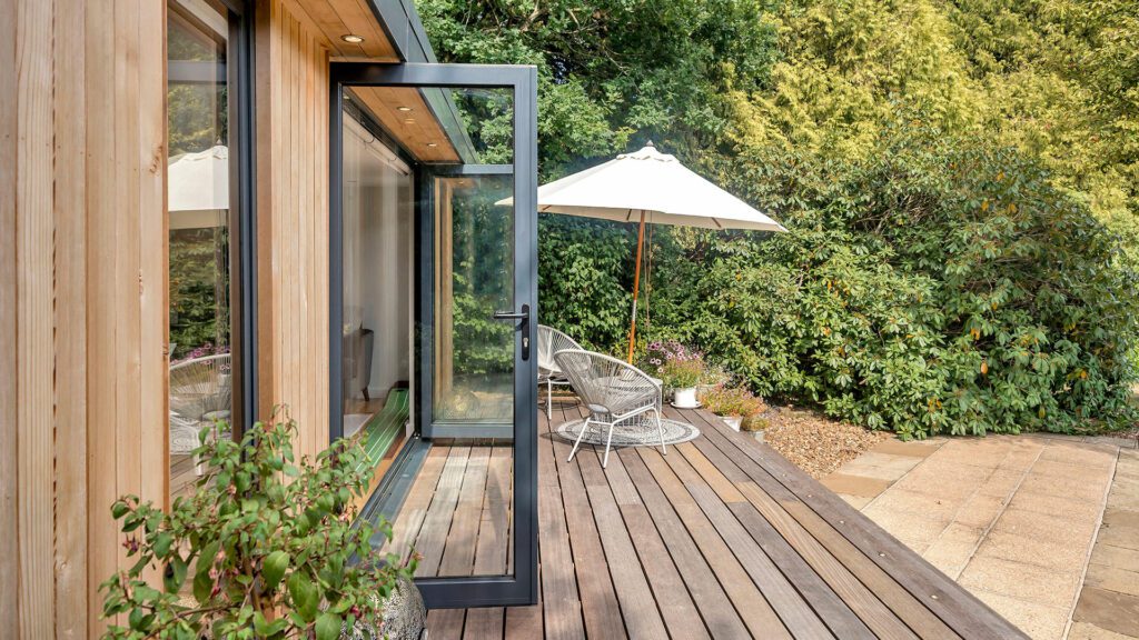 A wooden deck with a partially open sliding glass door, a white patio umbrella, wicker chairs, and plants, adjacent to an insulated garden office and lush garden.