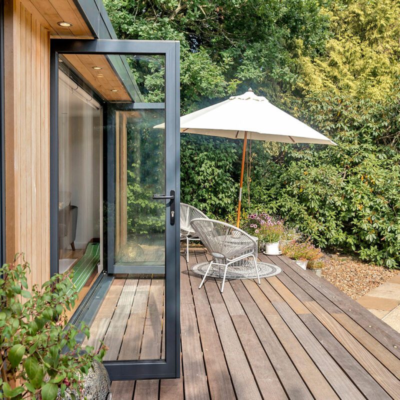 A wooden deck with a partially open sliding glass door, a white patio umbrella, wicker chairs, and plants, adjacent to an insulated garden office and lush garden.