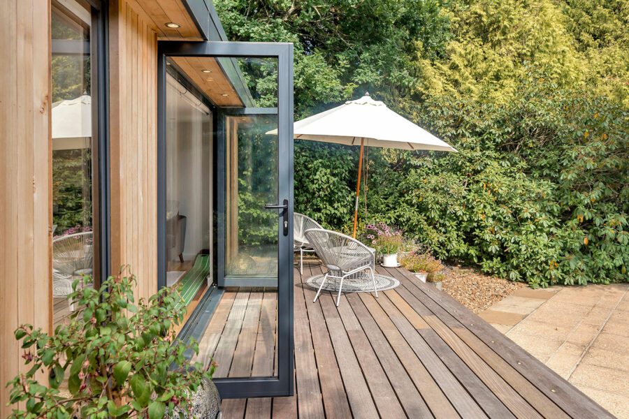 A wooden deck with a partially open sliding glass door, a white patio umbrella, wicker chairs, and plants, adjacent to an insulated garden office and lush garden.