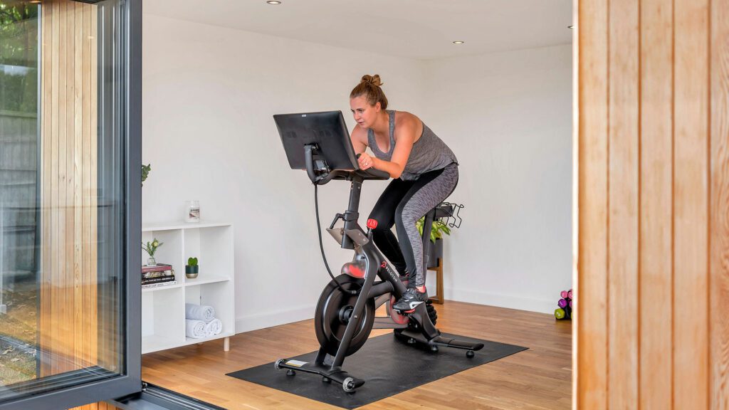 A person exercises on a stationary bike in a minimalist small garden room with a wooden floor, a white shelf, and a large glass door. The individual is focused on a screen attached to the bike.