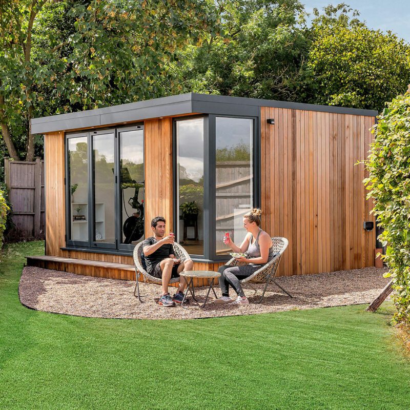 Two people sit outside a modern wooden cabin with large windows, surrounded by lush greenery and hedges, chatting and enjoying beverages after a refreshing workout in their nearby garden gym.